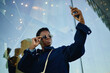 © TrueFrame Collective - Low angle shot of Black young adult woman holding smartphone and adjusting sunglasses while taking selfie outdoors, conveying mobile lifestyle and personal branding for social media marketing