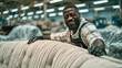 © pattozher - Craftsmanship and Pride: A skilled artisan, wearing work gloves and an apron, smiles warmly while working on a tufted sofa in a workshop. The background suggests a busy manufacturing environment.