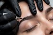 © Raisa - Cosmetologist works with brow microblading pen at beauty salon. Master in black gloves eyebrow tattoo correction for female client face. Pro permanent makeup artist uses tattoo machine tool for skin