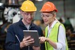 © Moopingz - Two industrial professionals wearing hard hats and safety goggles review data on a tablet in a factory.