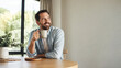 © Kowit - Man smiling while holding a cup of coffee, sitting at a wooden table near a window with natural light and indoor plants.