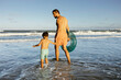 © kleberpicui - A father and young son walk hand in hand into gentle ocean waves, carrying an inflatable tube on a warm summer afternoon, capturing family bonding, relaxation and seaside fun.