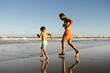 © kleberpicui - Father and son run and play along a sunny shoreline, barefoot on wet sand with reflections in shallow water. Warm light evokes joy, connection, health, and carefree summer family bonding at the beach.