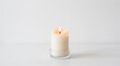 © SimplaStock - A single white candle burning brightly on a clear glass holder against a plain white background