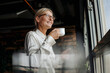 © Westend61 - Smiling businesswomanholding cup of coffee looking out of window