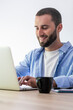 © simona - Young bearded man smiling and typing on a laptop, enjoying remote work and productivity with a cup of coffee on a light wooden desk against a white background