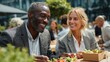© Alexei - African American man and Caucasian woman enjoying lunch outdoors at a table, surrounded by greenery and other people in a modern urban setting