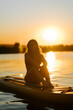 © Darius - Young woman sitting on a paddleboard during a golden sunset at a calm lake in the summer evening
