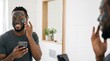 © Alexey - Happy African American man applying charcoal face mask while using smartphone in bathroom for male grooming and self care