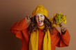 © Alliance - A woman in a yellow knit hat, orange sweater, and scarf playfully shouts with eyes closed, fingers in ears, holding autumn leaves. Against a brown background, she joyfully reacts to the fall season.