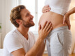 © Facundo - Happy future father smiling while gently touching his pregnant partner's belly in a bright home setting
