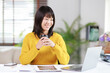© Matawut - Asian woman working from home, smiling and holding a coffee cup while using a laptop on a desk, representing remote work and freelance productivity