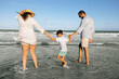 © kleberpicui - Parents walk hand in hand with their young son along a sunny shoreline, splashing in shallow ocean water and enjoying a relaxed summer family vacation filled with laughter and connection.