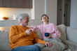 © DimaBerlin - Cheerful happy senior married couple sitting resting on couch at home joking and laughing together, joyful moment in elderly marriage, healthy positive relationships, man and woman talking and smiling