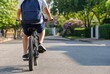 © Latarr - Schoolboy with blue backpack riding bicycle on sunny suburban street, back view of active child cycling through green neighborhood, healthy lifestyle and childhood independence concept.