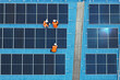 © unikyluckk - Aerial view of technicians installing solar panels on blue roof. Engineers maintaining solar energy system on a building rooftop. Top down view of workers in orange uniform fixing solar panels.