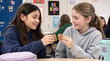 © ChaoticDesignStudio - Elementary students sharing chocolate chip cookie during lunch break in classroom. Two preteen girls sitting at desk and smiling while breaking snack in half.