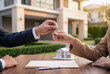 © Светлана Волынкина - Close-up of a house key being handed to a new owner over real estate documents and a home model. Property purchase, ownership, investment and residential housing concept