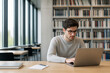 © Anastasiia - A young man wearing glasses sits at a wooden desk in a library, looking thoughtfully at his laptop.
