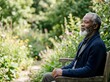 © Yuliia - Senior African American man with gray beard sitting on wooden bench in lush garden setting, wearing navy cardigan. Suitable for wellness, retirement, contemplation, and lifestyle editorial use.