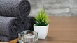 © sukanya - A minimalist still image set in a spa shows dark gray towels rolled up on a wooden tray, along with tealight candles and decorative potted plants.