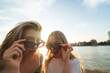 © pablo - Two young women peering over sunglasses, smiling at camera with river and golden hour sky