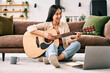 © HBS - Young woman playing guitar at home with laptop.