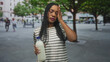 © Krakenimages.com - Woman holding a milk bottle by the cap with one hand, touching cheek and neck with the other, striped shirt and braids visible on a street plaza; quiet contemplation.