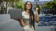 © Krakenimages.com - Teenage girl holding laptop with one hand, raises fist and smiles showing braces while looking at screen on city street plaza; achievement success joy.
