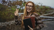 © Krakenimages.com - Redhead woman with glasses holding two books and flashing a peace sign by a stone wall in a forest; joy curious optimism.