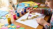 © Sangad - Child hand using brush to spread glitter on paper at a table with colorful craft supplies.