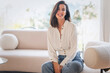 © gzorgz - Full length of a cheerful smiling confident woman sitting on the couch in front of a floor-to-ceiling window
