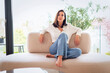 © gzorgz - Full length of a cheerful smiling confident woman sitting on the couch in front of a floor-to-ceiling window
