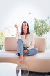 © gzorgz - Full length of a cheerful smiling confident woman sitting on the couch in front of a floor-to-ceiling window