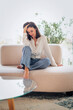 © gzorgz - Full length of a cheerful smiling confident woman sitting on the couch in front of a floor-to-ceiling window