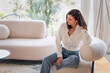 © gzorgz - Full length of a cheerful smiling confident woman sitting on the couch in front of a floor-to-ceiling window