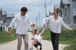 © ultramansk - Happy young family with little daughter cheer while their daughter leaps ahead on the road among modern new houses of a suburban housing estate, authentic everyday scene