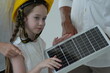 © ultramansk - Happy young family with little daughter in a toy hard hat let the girl in a toy helmet touch the photovoltaic cells up close in their future eco friendly house on a bright sunny day