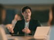 © Jiraphat - A man in a blazer is speaking and gesturing while seated at a table with a laptop, likely participating in a meeting or presentation.