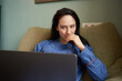 © Elena Kharichkina/Stocksy - Woman sits on couch while using laptop at home