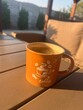 © GILPEDRO - Taza de café naranja con espuma sobre mesa de madera en una terraza durante el amanecer