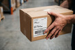 © Hani - Close-up of a person's hands holding a brown cardboard shipping box with a label and barcode in a warehouse setting.