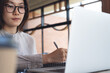© tippapatt - Asian business woman writing in notebook while working remotely on laptop at coffee shop. Freelancer planning work project, online working at cafe, freelance lifestyle concept
