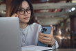 © tippapatt - Asian businesswoman using smartphone and working on laptop in cafe modern workspace, remote work, business casual, online business lifestyle, focused multitasking concept.