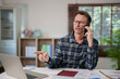 © Tj - Frustrated mature man having an angry conversation on a mobile phone, pointing finger at a laptop while working from home, expressing stress and problem solving issues