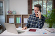 © Tj - Mature man working from home, talking on mobile phone while using laptop and analyzing business reports on desk, multitasking during a busy workday