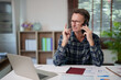 © Tj - Senior man communicating on smartphone while working remotely from home, multitasking with laptop and documents, managing business calls and finances in a casual setting