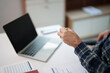 © Tj - Person working from a home office, holding a pen over business documents and analyzing data while using a laptop to manage tasks and achieve success