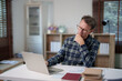 © Tj - Mature man wearing glasses and a plaid shirt, contemplating ideas and reviewing information on his laptop while working remotely at a desk in his home office