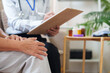 © Natee Meepian - Close up of senior patient hands resting while doctor takes notes on clipboard during consultation